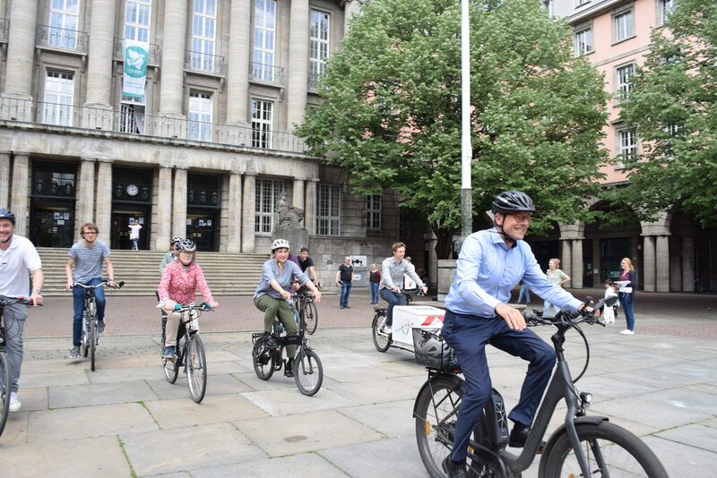 OB Uwe Schneidewind und weitere Menschen fahren vor dem Rathaus auf Fahrrädern los.