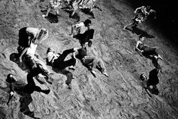 Szenenbild aus dem "Stück mit dem Schiff", die Tänzerinnen und Tänzer bewegen sich auf Sand und schiefer Bühnenebene