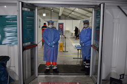 Eingang Impfzentrum: Zwei Mitarbeiter stehen in voller Schutzkleidung rechts und links der Tür.