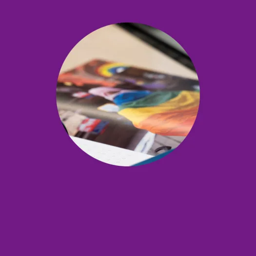 Eine Nahaufnahme eines aufgeschlagenen Magazins oder einer Broschüre, die ein Bild von Personen zeigt, die eine Regenbogenfahne tragen. Die Szene scheint Teil einer LGBTQ+-Veranstaltung oder Parade zu sein, wobei der Fokus auf der bunten Fahne und den beteiligten Personen liegt