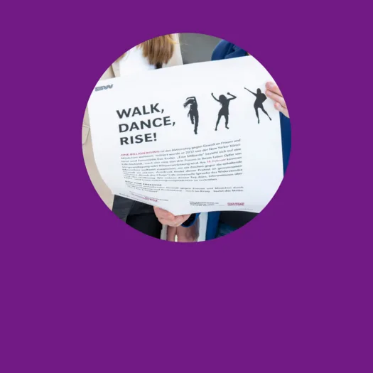 Zwei Personen halten ein Plakat mit der Aufschrift 'WALK, DANCE, RISE!' Auf dem Plakat sind Silhouetten von tanzenden Menschen abgebildet. Darunter befindet sich ein Text, der die Aktion 'ONE BILLION RISING' beschreibt, eine weltweite Bewegung gegen Gewalt an Frauen und Mädchen. Der Text erklärt, dass die Aktion 2012 ins Leben gerufen wurde und am 14. Februar weltweit stattfindet, um auf das Thema aufmerksam zu machen und Solidarität zu zeigen.