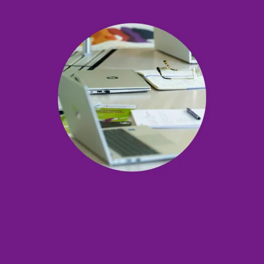 Ein Tisch in einem Büro oder Besprechungsraum, auf dem mehrere Laptops, Notizblöcke, Stifte und Dokumente liegen. Eine Brille liegt auf einem offenen Notizbuch, und im Hintergrund sind bunte Poster oder Plakate zu erkennen.