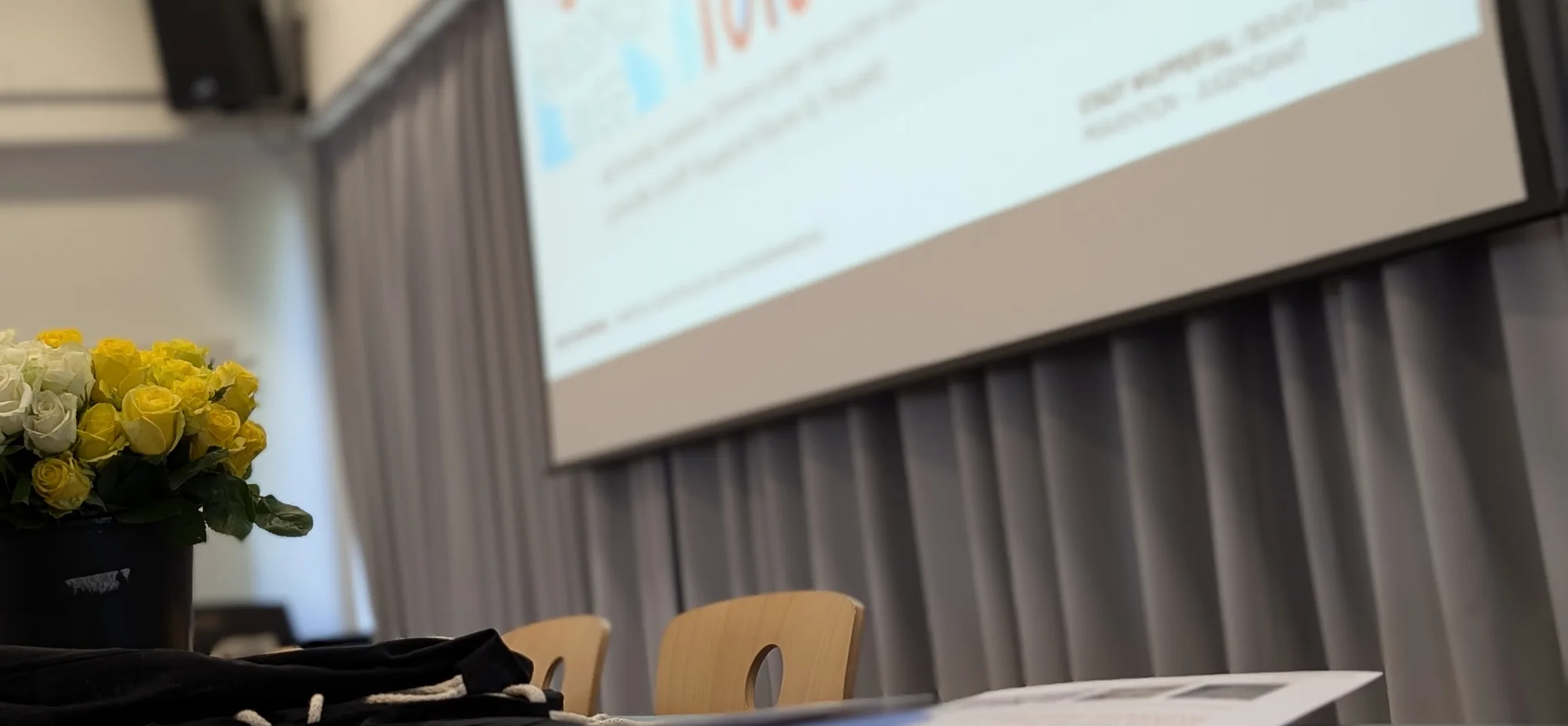 Schulraum mit Leinwand, auf der eine Präsentationsfolie zum Thema Toleranz und Vielfalt gezeigt wird. Im Vordergrund liegen auf einem Tisch Tagungsmaterialien und Blumen