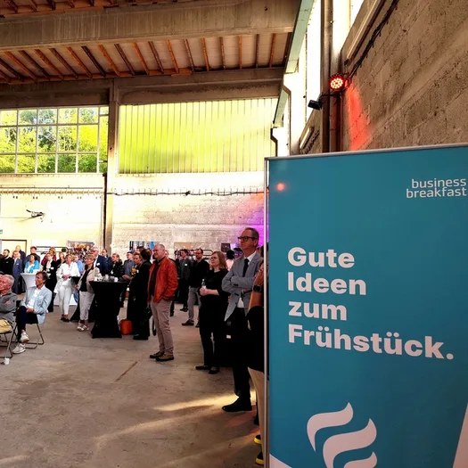 Vorne: Business Breakfast Plakat. Hinten: Gäste in der alten Glaserei