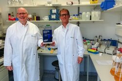 Die beiden Geschäftsführer von AiCuris stehen im Labor und halten den Rheinland Genial Preis zwischen sich.