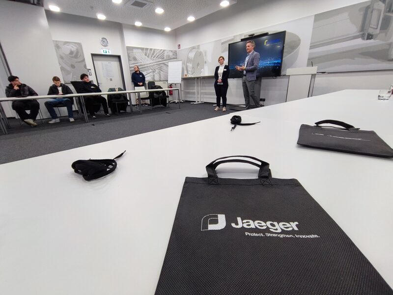 Tasche mit Jaeger-Logo im Vordergrund, im Hintergrund Schüler und Unternehmensvertreter