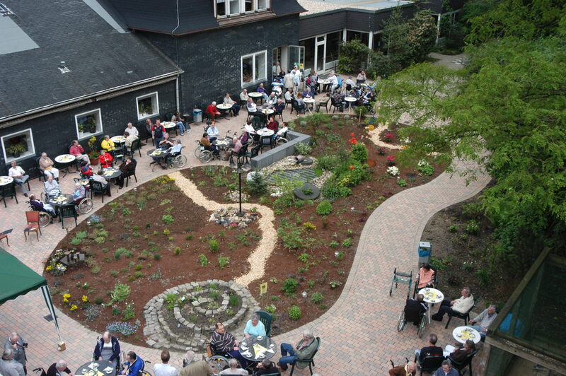 Ansicht Gartenfläche mit Kräuterspirale, außen Bestuhlung mit Bewohnerinnen