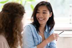 Asian woman talking with colleague sitting at desk indoors