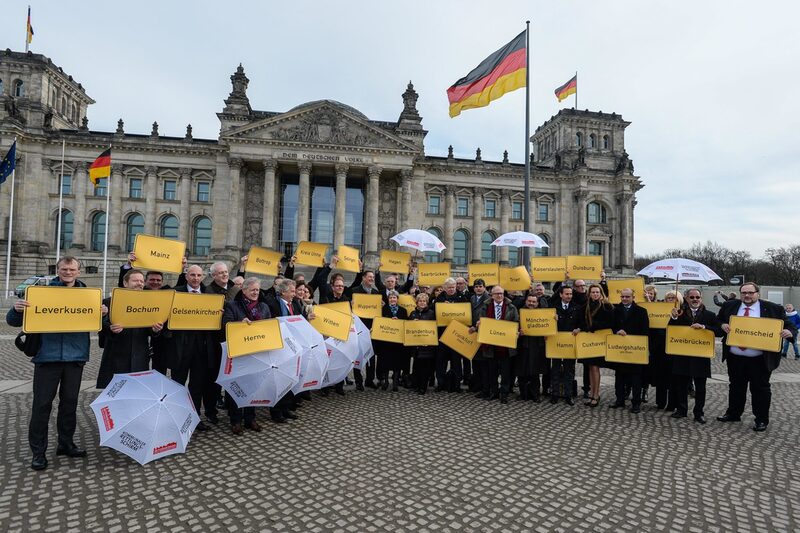 Die Oberbürgermeister und Kämmerer der Aktionsstädte stehen in Berlin vor dem Reichstag und halten Schilder mit den Namen ihrer Kommunen sowie weiße Regen-"Rettungs"-Schirme hoch.