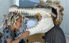 Ein kleines Mädchen im Profil albert mit einer Krokodilmaske herum, die eine Erwachsener auf dem Kopf trägt.