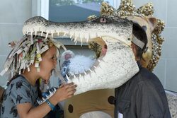 Ein kleines Mädchen im Profil albert mit einer Krokodilmaske herum, die eine Erwachsener auf dem Kopf trägt.