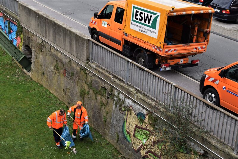 Zwei Mitarbeiter des Eigenbetriebs Straßenreinigung sammeln Müll ein an der Schwebebahnstation Adlerbrücke