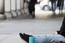 Ein Mensch sitzt in der Fußgängerzone am Boden. Zusehen sind nur die Beine und eine blaue Tasse.