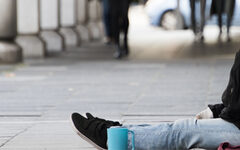 Ein Mensch sitzt in der Fußgängerzone am Boden. Zusehen sind nur die Beine und eine blaue Tasse.