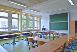 Blick in ein leeres Klassenzimmer mit Tafel, Tischen, Stühlen und Lehrerpult
