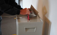 Hände, die einen Umschlag in eine Wahlurne stecken
