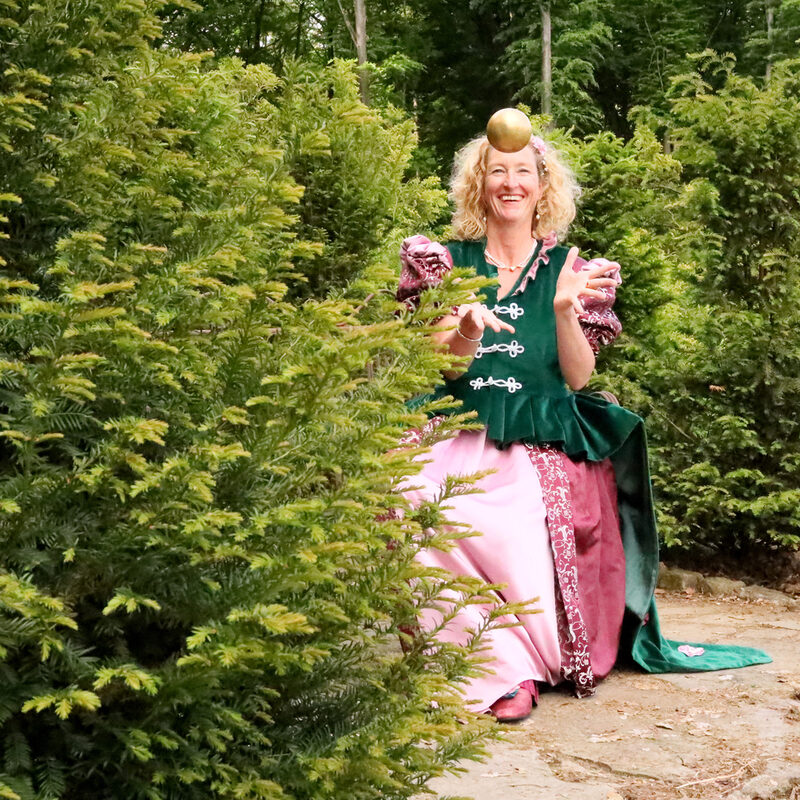 Märchenerzählerin Miss Fairytail sitzt im historischen Kostüm zwischen Bäumen und spielt mit einem goldenen Ball
