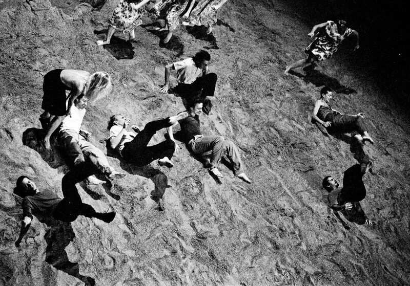Szenenbild aus dem "Stück mit dem Schiff", die Tänzerinnen und Tänzer bewegen sich auf Sand und schiefer Bühnenebene