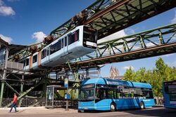 Ein blauer Wasserstoffbus der Stadtwerke steht unter dem Schwebebahngerüst, an dem eine himmelblaue Schwebebahn fährt