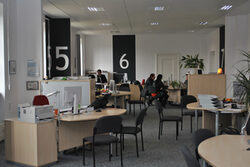 Großraumbüro mit Schreibtischen und Stühlen, im Hintergrund Mitarbeiter und Besucher