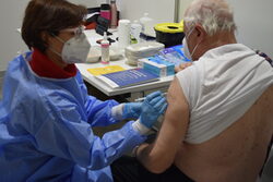 Ärztin in blauem Kittel und Maske impft älteren Patienten
