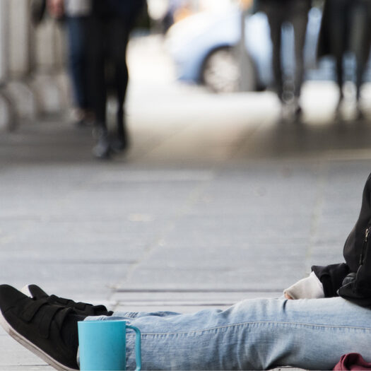 Symbolbild: Mensch sitzt in der Fußgängerzone auf Steinboden, Tasse neben sich