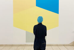 Die Künsterlerin Franziska Holstein, von hinten zu sehen, betrachtet ihr Bild in klaren geometrischen Formen und den Farben gelb, grün und blau im Von der Heydt-Museum