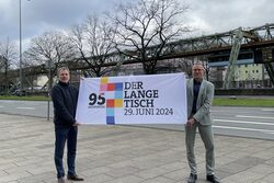 WMG-Chef Martin Bang und OB Uwe Schneidewind halten ein Plakat mit der Aufschrift "Der Lange Tisch" hoch, sie stehen dabei an der B7. Im Hintergrund fährt eine Schwebebahn.