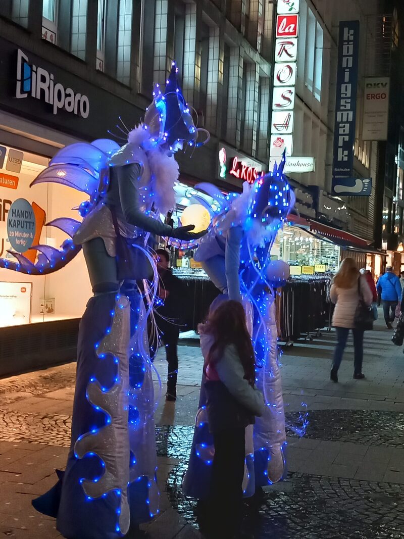 zwei Künstlerinnen in blauen Leuchtkostümen in der Fußgängerzone