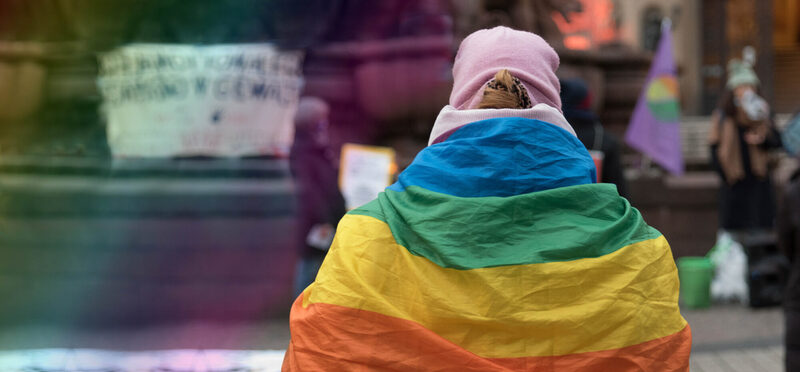 Eine Person steht mit einer Regenbogenfahne umgehangen mit dem Rücken zum Betrachter.