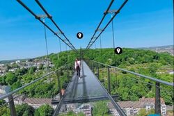 Screenshot zeigt virtuelle Sicht auf und von der geplanten Hängebrücke. Eine Frau läuft etwas weiter vorne, unter der Brücke sind Gebäude und Bäume zu sehen.