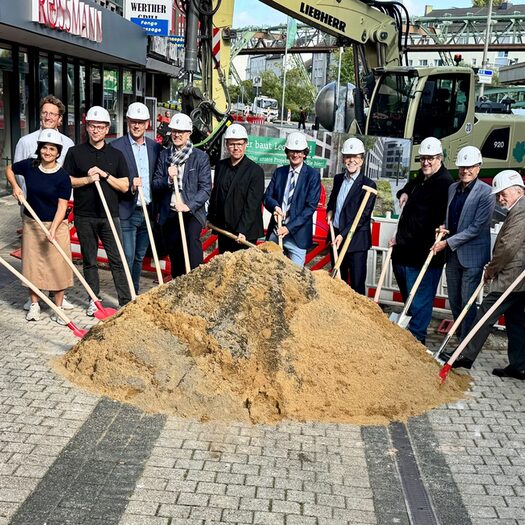 Die am Projekt Beteiligten halten Schaufeln in den Händen, die sie in Richtung eines Sandhaufens halten.