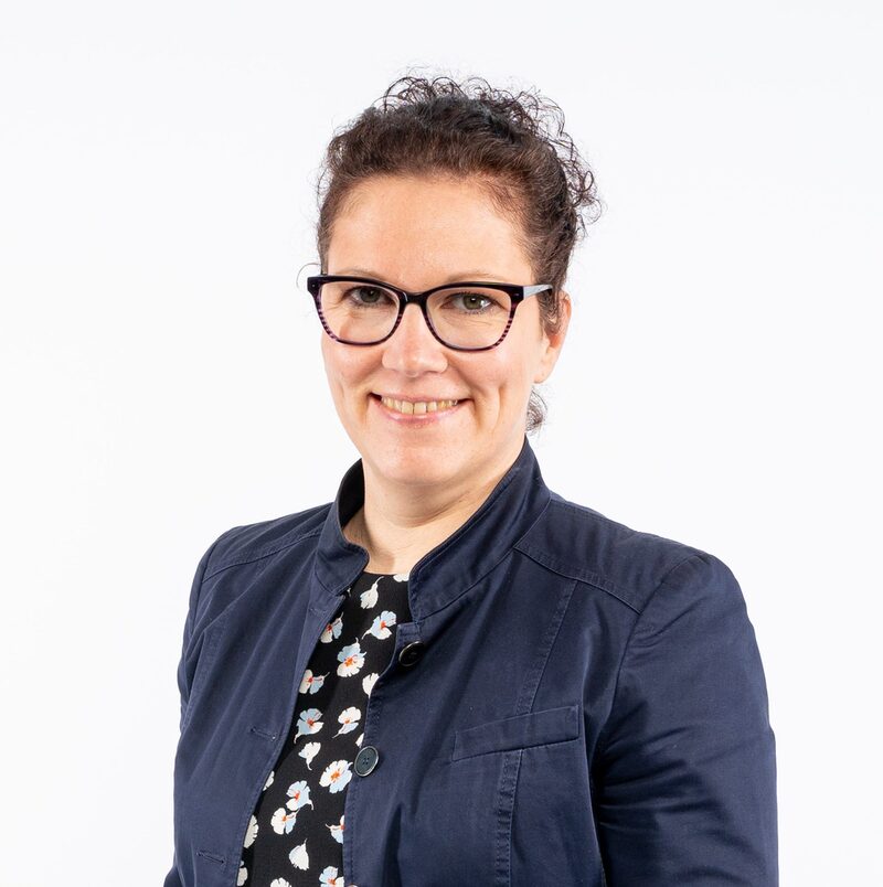 Zu sehen ist Sandra Heinen. Sie ist die Behindertenbeauftragte der Stadt Wuppertal. Sie hat braune lockige Haare, die sie in einem Dutt zusammengebunden hat. Sie trägt eine Brille und einen blauen Blazer. Sie lächelt und blickt in die Kamera.